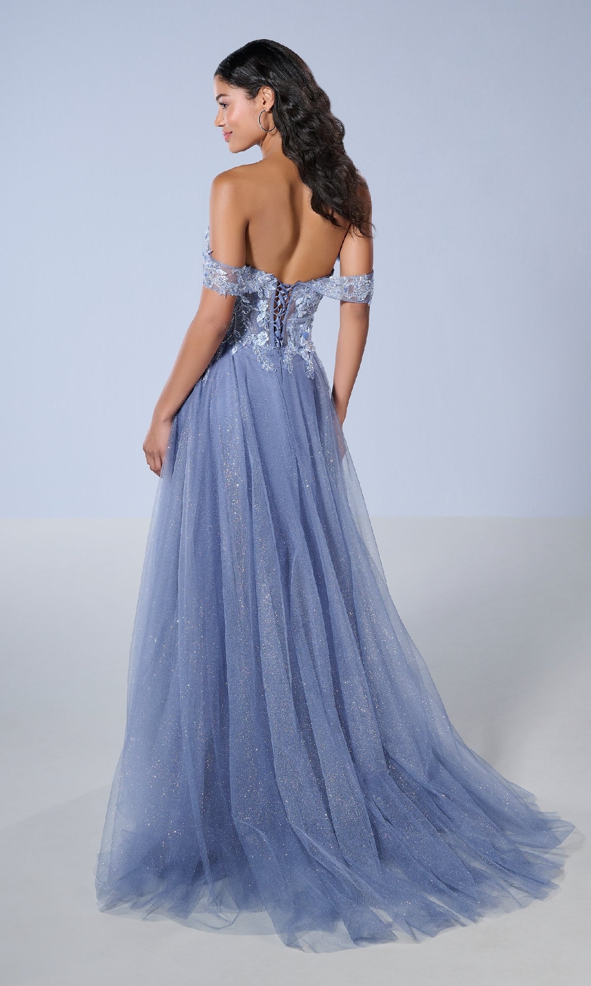 A woman stands elegantly against a plain light background wearing the Tiffany 16243 long prom dress, an off-the-shoulder lavender gown with a floor-length glitter tulle skirt and lace-up back.